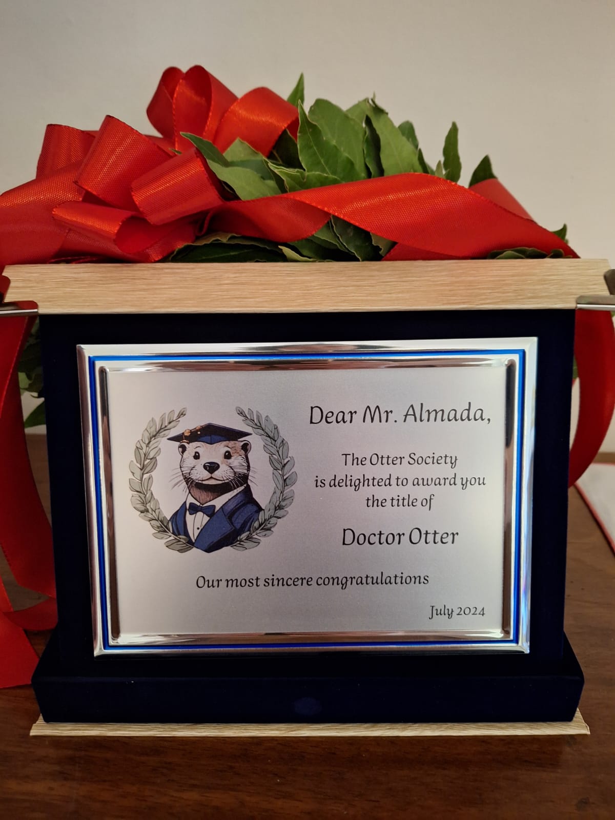 A commemorative plaque, featuring an otter in black tie and a graduation hat. The plaque says: "Dear Mr Almada,
The Otter Society is delighted to award you the title of
Doctor Otter
Our most sincere congratulations"