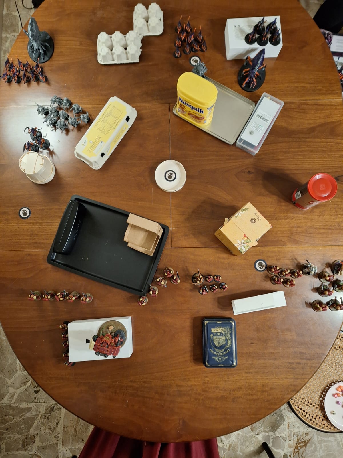 A table in which a game of Warhammer 40k is being played. The table is full of cans, boxes, and other household items being used as terrain. The armies are a mostly-painted Tyranid army and a fully-painted group of Blood Ravens Space Marines.